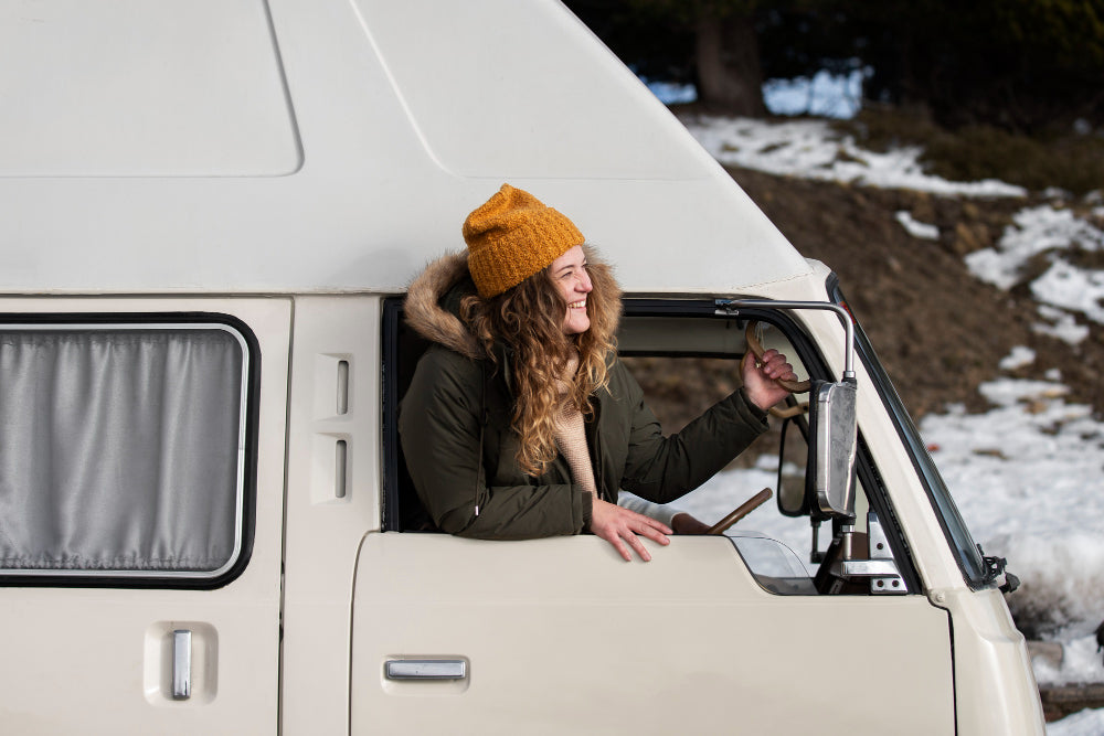 Person with curly hair wearing a yellow beanie and green jacket leaning out of a car window in a snowy landscape.