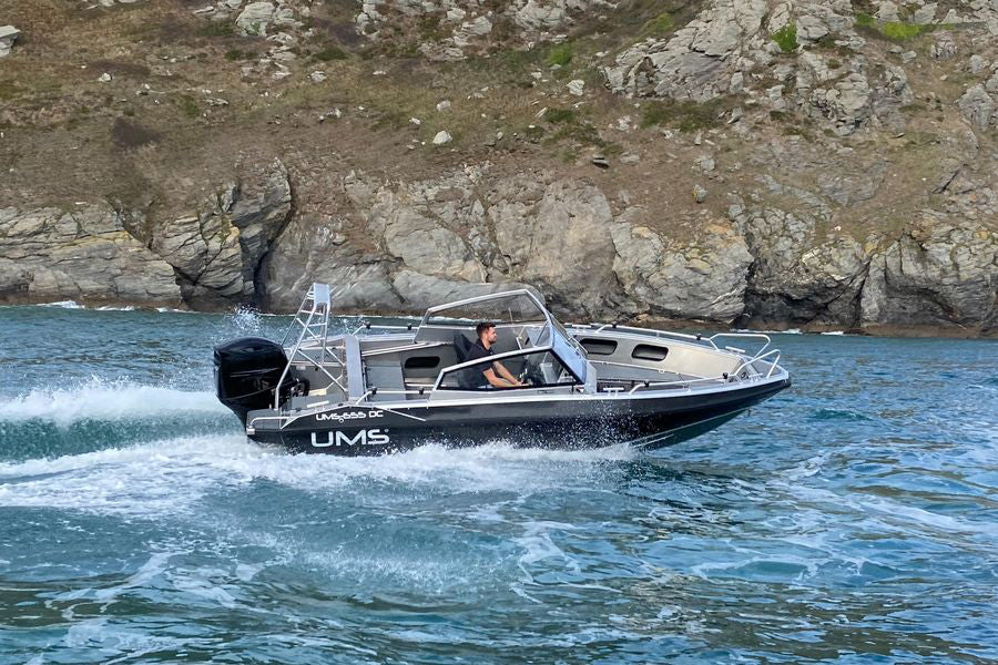 A man steering the UMS 655 DC aluminum boat at speed near a rocky shoreline.