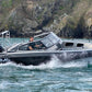 A man piloting the UMS 655 DC aluminum boat at speed near a rocky coastline.