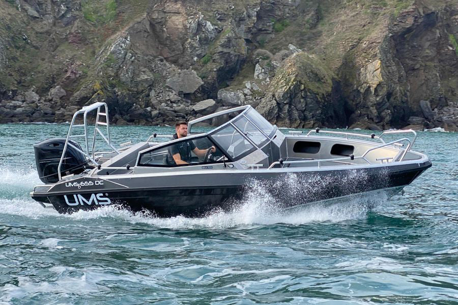A man piloting the UMS 655 DC aluminum boat at speed near a rocky coastline.