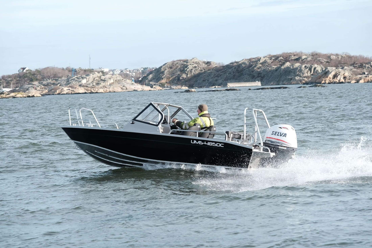 A person is driving an Aluminum Boat UMS 485 DC on open water near rocky shores, showcasing the boat's performance and design.